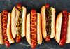 Hot Dog: Sokak Lezzetlerinin Vazgeçilmezi Dünya mutfağında geniş bir yer edinen hot dog, özellikle Amerika Birleşik Devletleri’nde adeta bir sembol haline gelmiştir. Kökeni 19. yüzyılın sonlarına kadar uzanan bu yiyecek, Almanya’dan Amerika’ya göç edenlerin beraberinde getirdiği sosis kültürü ile ortaya çıkmıştır. Uzun ekmek arasına yerleştirilen sosisin seneler içinde farklı soslarla buluşması, hot dog’u bugünkü popüler formuna kavuşturmuştur. Konulu bir haber görseli.