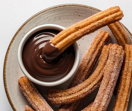 Churros, son yıllarda yalnızca İspanya’nın geleneksel sokak lezzeti olmaktan çıkıp dünya genelinde kafe menülerinin favorisi haline geldi. Özellikle Türkiye’de tatlı kültürünün giderek çeşitlenmesiyle birlikte churros popüler bir alternatif olarak öne çıkıyor. Çıtır yapısı, şeker ve tarçınla kaplanan dış yüzeyi ve sıcak çikolata sosuyla birleştiğinde ortaya çıkan benzersiz tat, birçok insanın yeni gözdesi haline gelmiş durumda. Konulu bir haber görseli.