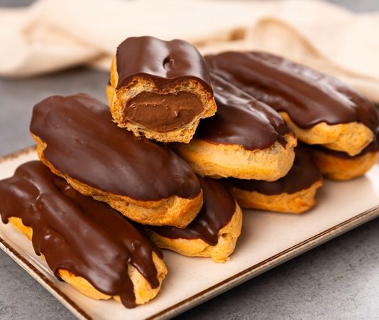 Fransız pastacılığının dünya mutfağına kazandırdığı en özel tatlardan biri olan eclair, yalnızca görünümüyle değil, ince dokusu ve hafif dolgusu ile de tatlı severlerin vazgeçilmezi olarak gösteriliyor. Günümüzde pek çok pastane menüsünde rastladığımız bu tatlı, aslında aristokrat mutfaklarından çıkıp modern mutfaklara taşınan çok katmanlı bir tarihe sahip. Özellikle çikolata kaplaması ve iç dolgusu ile bilinen eclair, Avrupa’da uzun yıllardır tüketilen bir tatlı olmakla birlikte, son yıllarda Türkiye’de de giderek daha fazla tercih edilmeye başladı. Pastacılığın gelişimi ile beraber farklı dolgular, farklı çikolata türleri ve daha çeşitli glazürlerle sunulan bu tatlı, gastronomik açıdan ciddi bir çeşitliliğin de öncüsü haline geldi. Konulu bir haber görseli.