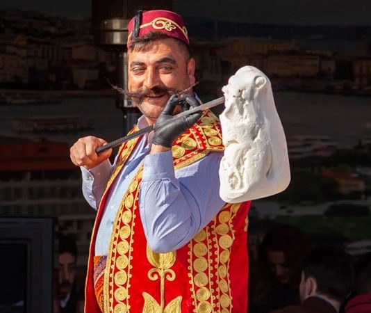 Gelenekten Lezzete Maraş Dondurmasının Hikâyesi ve Günümüzdeki Yeri Türkiye’nin gastronomi mirası içinde kendine özgü yapısı, üretim tekniği ve kültürel arka planıyla öne çıkan Maraş dondurması, yalnızca bir tatlı değil aynı zamanda köklü bir geleneğin günümüze taşınmış hâli olarak kabul ediliyor. Özellikle yaz aylarında yoğun ilgi gören bu eşsiz lezzet, dayanıklı yapısı, bıçakla kesilebilen kıvamı ve kendine has aromasıyla klasik dondurmalardan ayrılıyor. Kahramanmaraş ile özdeşleşen bu ürün, bugün hem yerel ekonominin hem de Türkiye’nin tanıtım yüzlerinden biri hâline gelmiş durumda. Konulu bir haber görseli.